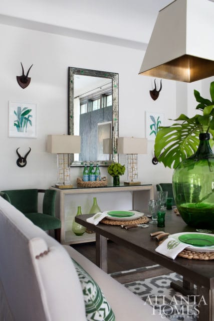 Dining room with dark wood table set with green plates and white napkins, a large green glass vase with monstera leaves, and a console table with lamps and decorative items.