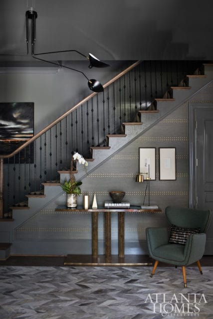 Gray entryway with a staircase featuring black spindles and a wood handrail, patterned wallpaper, a console table with decor, and a green armchair.