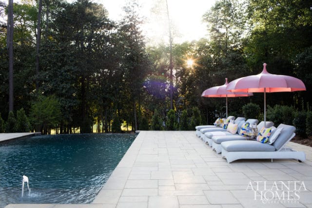 Outdoor pool area with light stone patio, lounge chairs with pink umbrellas, and lush green trees in the background.