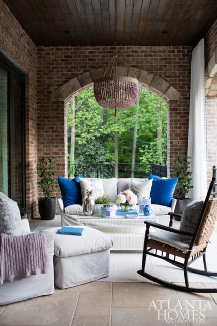 Covered porch with brick walls, wood-paneled ceiling, and arched opening revealing lush greenery. Furnished with a white sofa, armchair, rocking chair, and coffee table adorned with blue and white decor.
