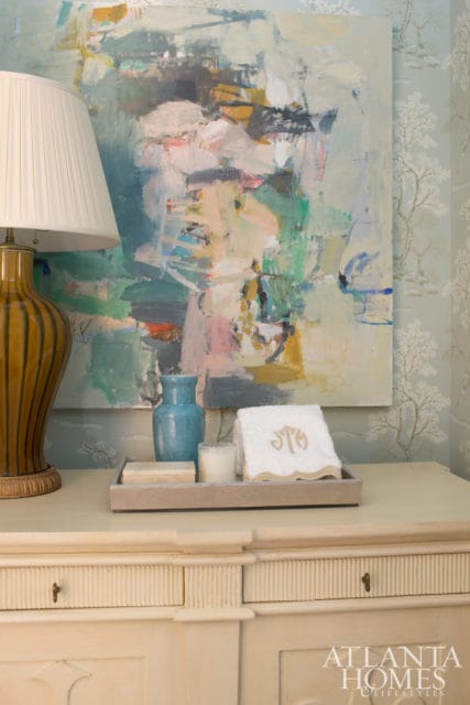 Cream dresser with fluted drawers topped with a tray holding a blue vase, candle, and monogrammed towel, beneath an abstract painting and tree-patterned wallpaper.