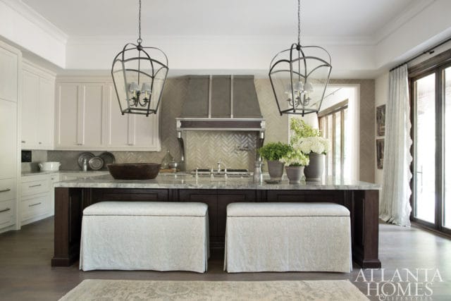Kitchen island with dark wood base, marble countertop, and upholstered benches. White cabinetry, herringbone tile backsplash, and large lantern pendant lights are visible.