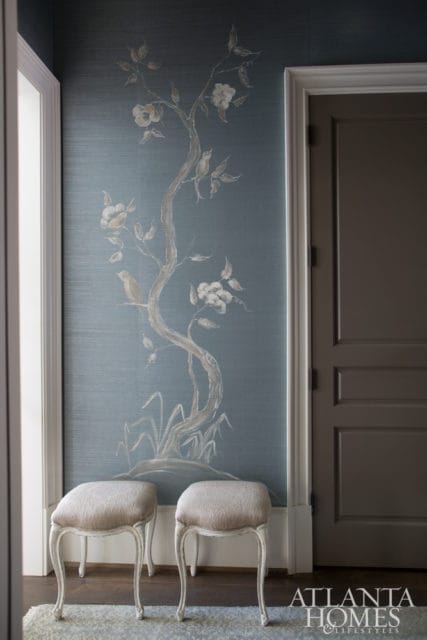 Hallway with blue grasscloth wallpaper featuring a hand-painted chinoiserie tree and birds, flanked by a white door frame and a dark gray door, with two upholstered stools in the foreground.