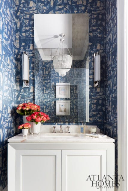 Powder room with blue abstract wallpaper, white vanity, marble countertop, crystal chandelier, and floral arrangements.