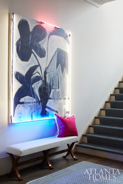 Hallway with abstract blue and white floral art illuminated by neon lights, a white bench with a pink pillow, and a carpeted staircase.