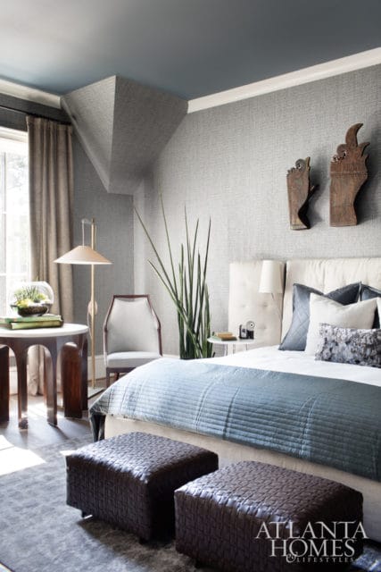 Bedroom with textured gray wallpaper, a blue quilted bedspread, tufted headboard, and two dark brown woven ottomans.