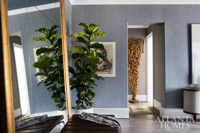 Hallway with blue patterned wallpaper, a large mirror reflecting a fiddle leaf fig plant, a brown leather bench with a throw blanket, and a doorway revealing a sculpture made of woven material.