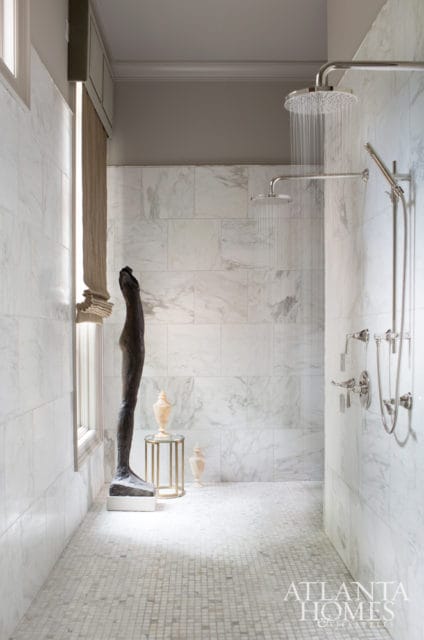 Modern bathroom with marble tile walls and floor, featuring a large walk-in shower with dual showerheads and chrome fixtures. A tall, dark sculpture stands near a window.