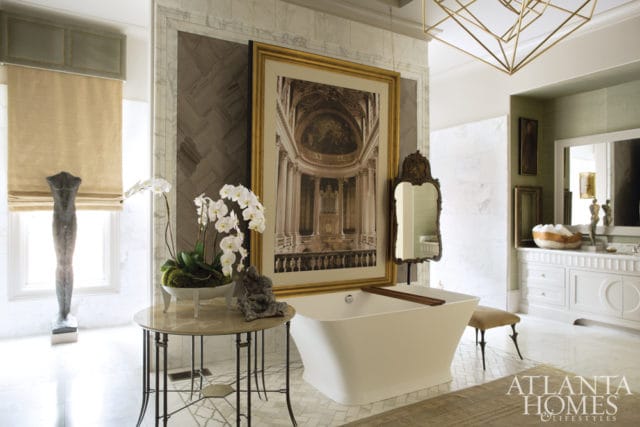 Modern white freestanding bathtub in a luxurious bathroom with marble floors, a large framed artwork of a cathedral, and orchids on a side table.