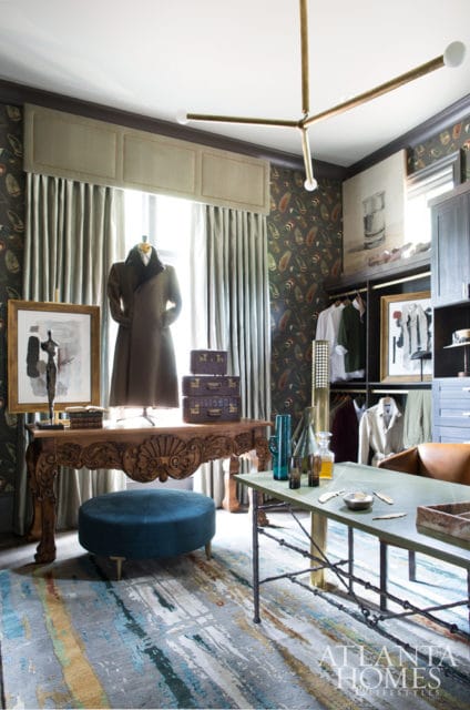 Dressing room with dark patterned wallpaper, a mannequin in a brown coat, ornate wooden desk, teal ottoman, and a walk-in closet with clothing.