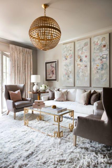 Living room with a white sofa, plush white rug, gray wingback chairs, a gold Greek key coffee table, and abstract triptych art on the wall.