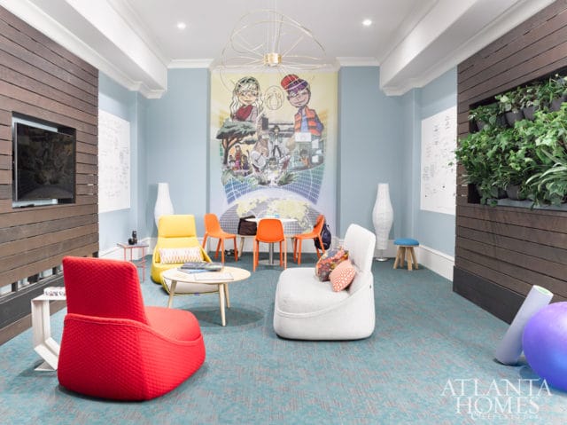 Light blue room with a large mural of children and a world map, featuring red and yellow armchairs, white beanbag chairs, orange chairs, and a vertical garden wall.