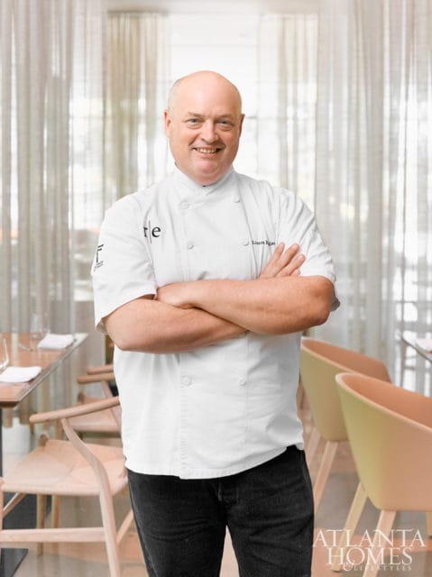 Chef Linton Hopkins smiles with arms crossed in a restaurant dining room, wearing a white chef's coat and dark jeans.