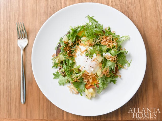 White plate with a salad topped with a poached egg and crispy fried onions, next to a silver fork on a wooden table. Atlanta Homes & Lifestyles logo in the bottom right corner.