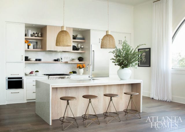 Modern white kitchen with a light wood island, four industrial-style stools, woven pendant lights, and open shelving displaying decor.