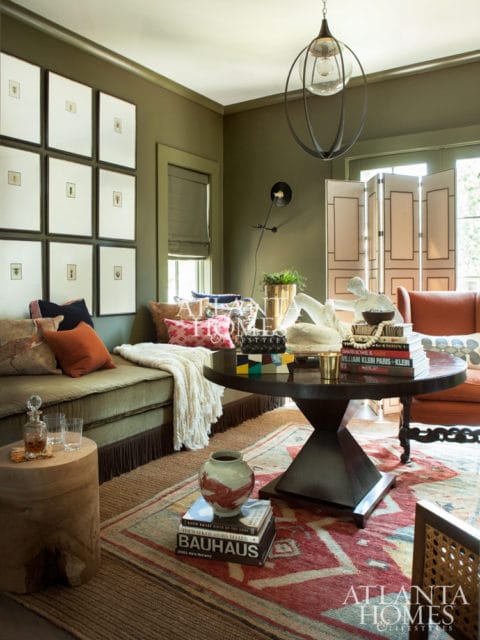 Olive green living room with a sectional sofa, gallery wall of botanical prints, round dark wood coffee table with books, and a patterned rug.