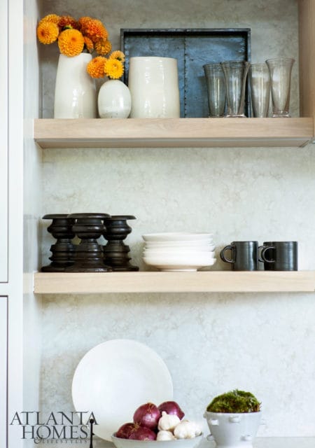 White vases with orange flowers, dark tray, and clear glasses on a wooden shelf above dark candle holders, stacked white bowls, and black mugs. Below are a white plate, bowl of red onions and garlic, and a moss-filled planter.