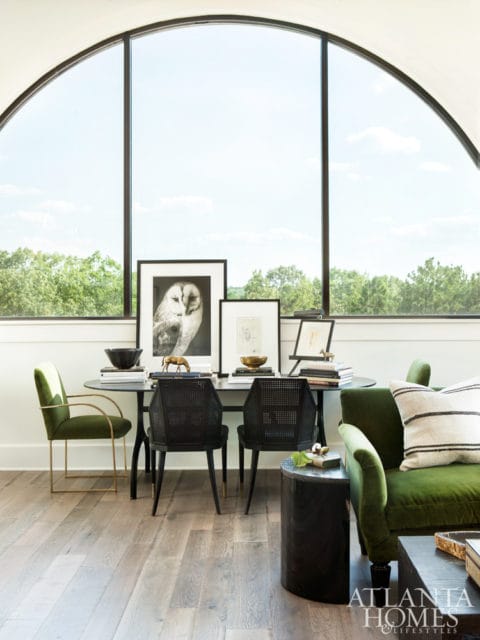 Living room with a large arched window overlooking trees, featuring a green velvet sofa, black cane-backed chairs, and a small round table with framed art.
