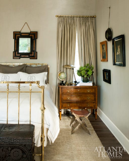 Bedroom with brass bed, white bedding with monograms, wooden dresser, globe, and taupe curtains.