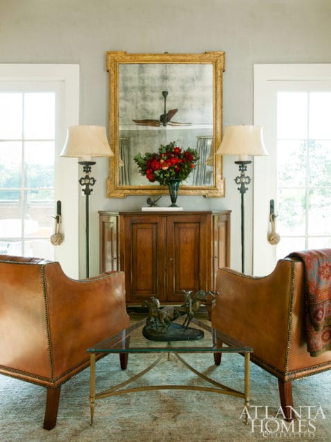 Living room with two brown leather armchairs flanking a wooden cabinet topped with a large antique mirror and a vase of red flowers.