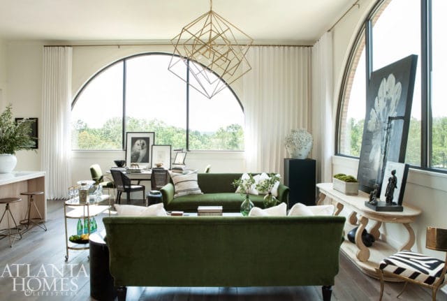 Living room with two green velvet sofas, a gold geometric chandelier, and a large arched window overlooking trees.