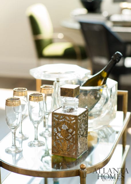A gold-embossed decanter sits on a glass bar cart next to champagne flutes and a bottle of champagne in an ice bucket.