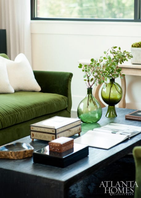 Green velvet sofa with white pillows next to a dark wood coffee table holding decorative boxes, green glass vases with branches, and an open book.