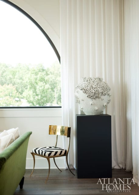 Living room with a green sofa, a gold-framed chair with zebra print upholstery, and a white textured vase on a black pedestal, with floor-to-ceiling white curtains and an arched window overlooking trees.