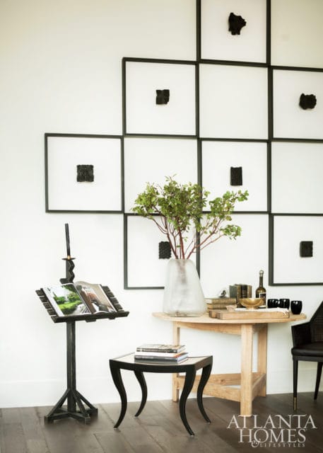 Modern living room with a grid of framed abstract art, a music stand with an open book, a small black side table with books, and a round wooden table with a vase of greenery, books, and glassware.