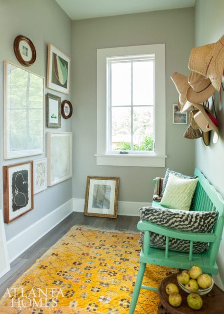 Entryway with pale gray walls, a mint green bench with patterned pillows, a yellow patterned rug, and a collection of framed art and straw hats.