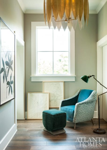 Hallway with sage green walls, a large gold leaf chandelier, a blue armchair with a leopard print accent, a green ottoman, and framed botanical sketches.