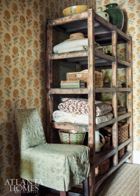 A rustic wooden shelf unit filled with folded blankets, books, baskets, and decorative vases, next to a slipcovered chair in a room with floral wallpaper.