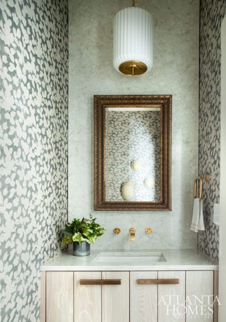 Powder room with patterned wallpaper, marble countertop, light wood vanity with brass hardware, and a gold-framed mirror above the sink.
