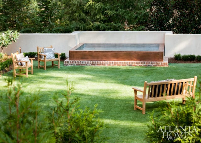 Backyard with a copper-clad hot tub, brick base, and surrounding green lawn. Two wooden chairs with patterned pillows and a wooden bench with dark cushions are placed on the grass.