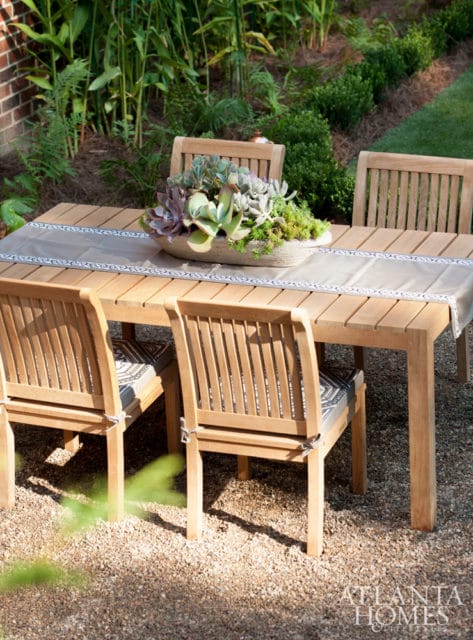 Wooden dining table and chairs set on gravel patio with succulent centerpiece and lush green landscaping in the background