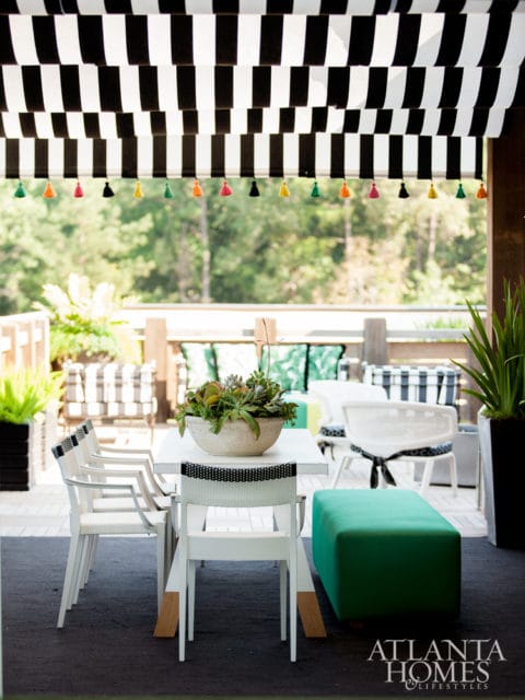 Outdoor dining area with a white table and chairs, a green ottoman, and a black and white striped awning decorated with colorful tassels.