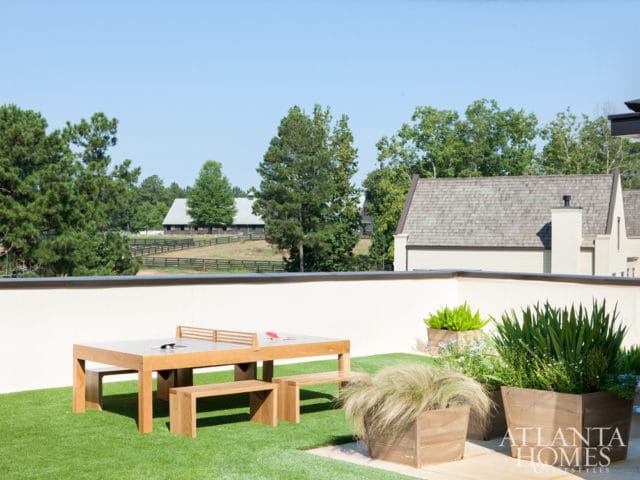 Rooftop patio with artificial turf, a wooden ping pong table and benches, and large wooden planters filled with greenery. In the background, a barn and house are visible amidst trees.