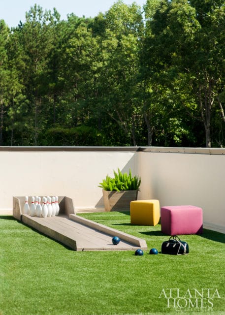 Outdoor bowling alley with white pins and blue balls on artificial turf, next to a planter and colorful ottomans.