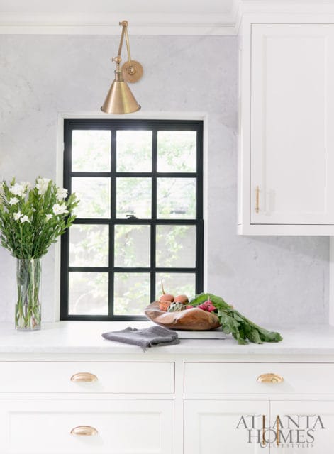 White kitchen with marble backsplash, black grid window, brass swing arm sconce, and white cabinetry with brass hardware.