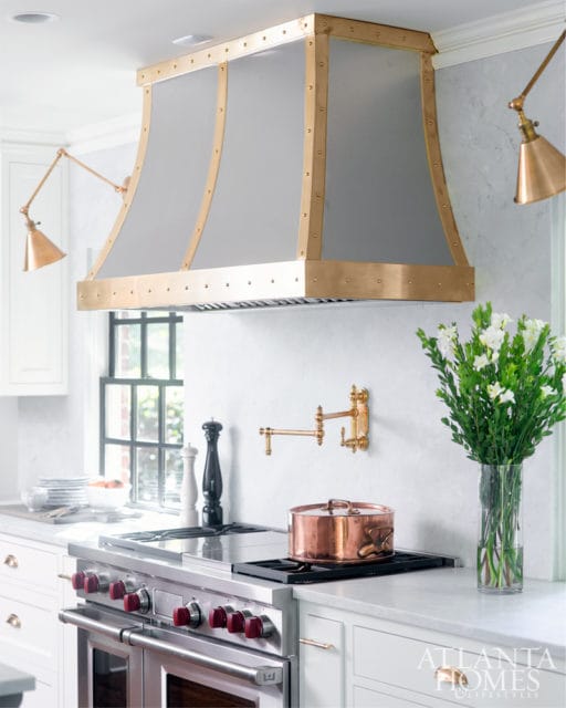 White marble kitchen with a large stainless steel range, copper pot, and a custom range hood with brass accents.