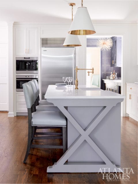 Modern kitchen with a light gray island featuring X-frame supports, white quartz countertop, and brass pot filler. White cabinetry, stainless steel appliances, and light gray upholstered bar stools are visible.