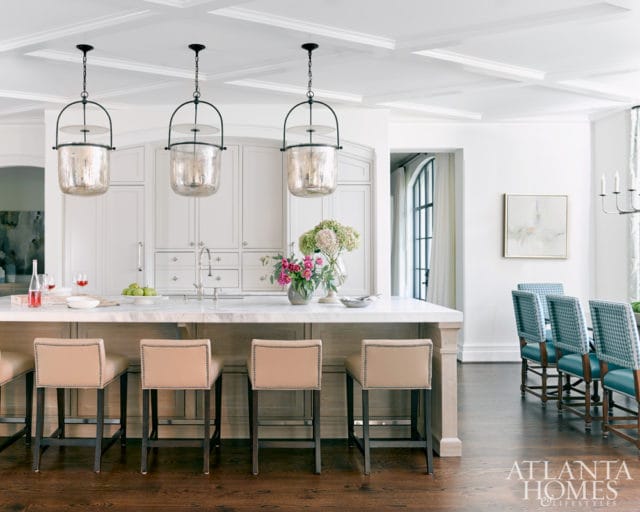 Kitchen island with marble countertop and tan upholstered stools, facing white custom cabinetry with stainless steel appliances. Three mercury glass pendant lights hang above.