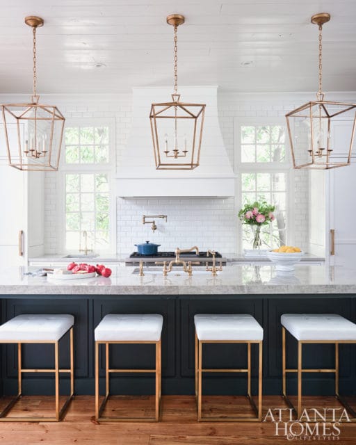 White subway tile backsplash and range hood in a kitchen with a large marble island topped with gold faucets and white bar stools.