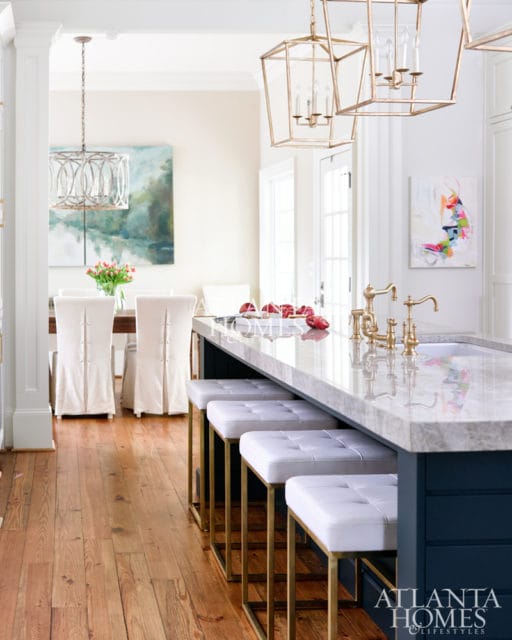 Kitchen island with white upholstered stools and marble countertop, brass faucets, and navy blue cabinetry. Wood floors and dining area visible in background.