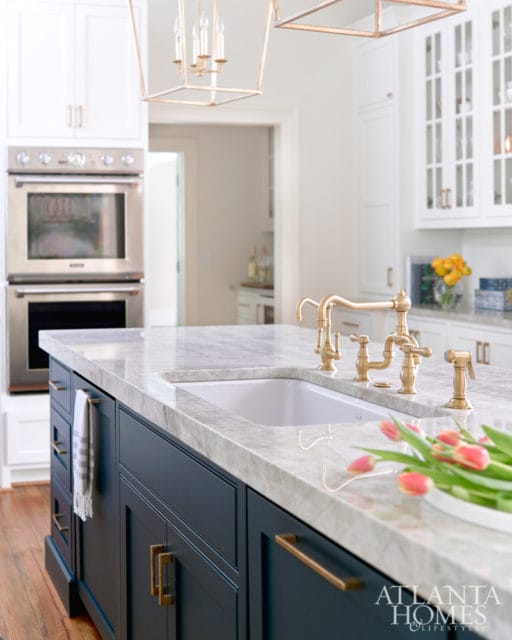 Dark blue kitchen island with marble countertop and brass faucet, white cabinetry with double ovens in background, Atlanta Homes & Lifestyles logo in corner
