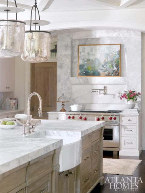 Kitchen island with marble countertop and farmhouse sink, stainless steel range with red knobs, and abstract art above the range.