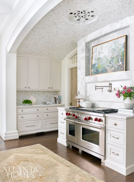 White kitchen with a mosaic tile ceiling, marble backsplash, stainless steel range with red knobs, and custom white cabinetry.