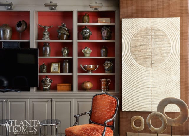 Built-in gray cabinetry with red shelves displaying various ceramic jugs, pottery, and decorative objects. An orange upholstered armchair sits in front of the shelves.
