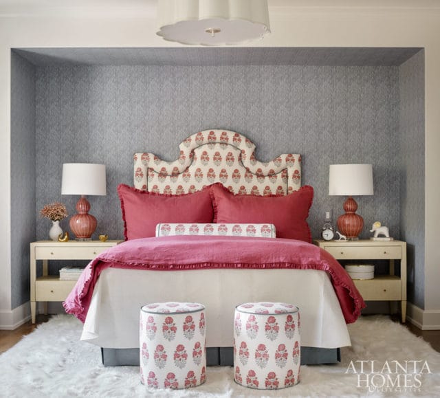 Bedroom with a gray patterned wallpapered alcove, a white bed with pink and red floral headboard, red pillows, and two white nightstands with coral lamps.