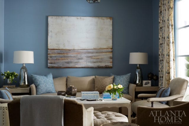 Living room with blue walls, a large abstract painting, beige sofa with blue patterned pillows, and a white coffee table with floral arrangement.